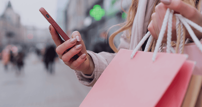 Consumer checks online deals on smartphone amid pink shopping bags and urban storefront backdrop