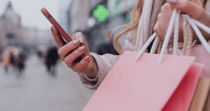 Consumer checks online deals on smartphone amid pink shopping bags and urban storefront backdrop